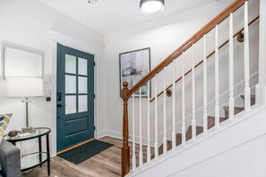 A hallway with a wooden staircase, a blue door, a small table with a lamp, and a framed picture on the wall.