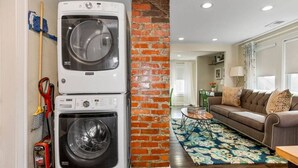 Un espace buanderie avec des machines à laver et des sèche-linge superposés, un mur en briques, un salon avec un canapé et un tapis à motifs, et un coin repas avec une table et des chaises.