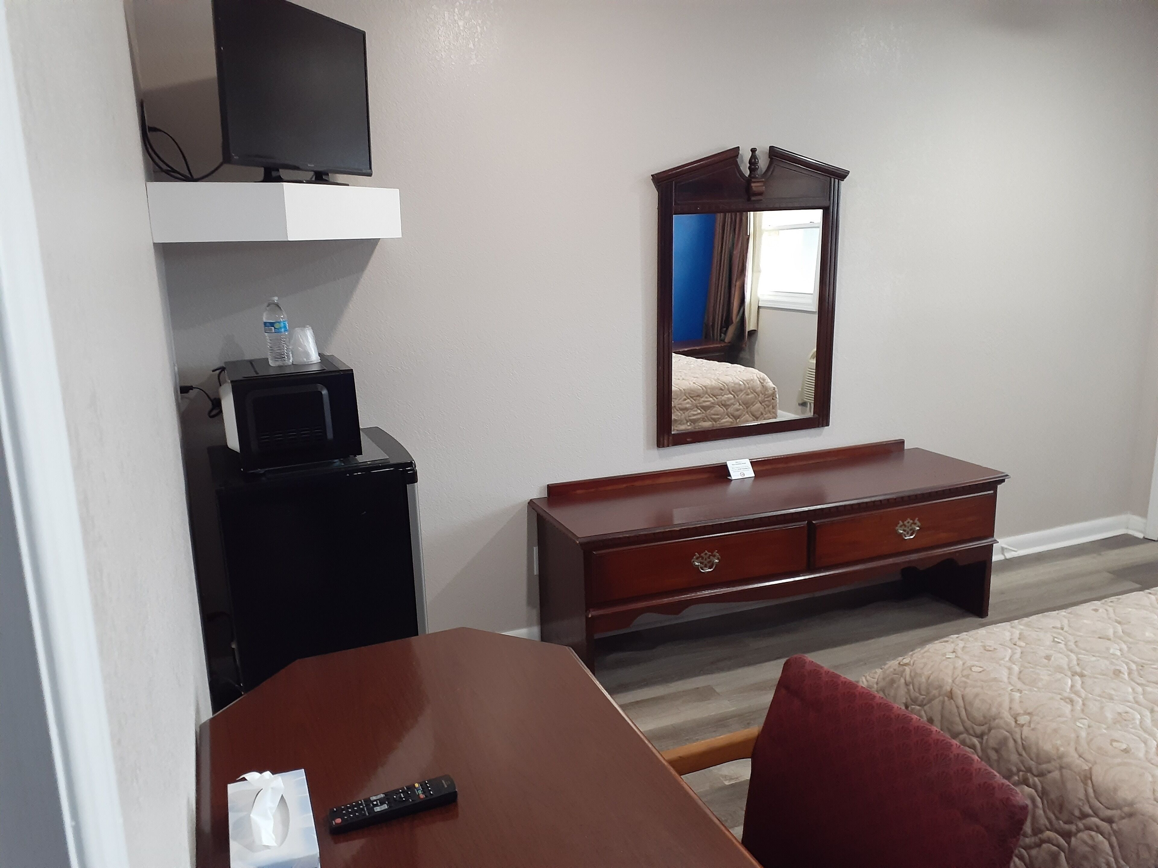 Quarto de hotel com cama, cômoda de madeira, televisão em uma estante e espelho na parede.
