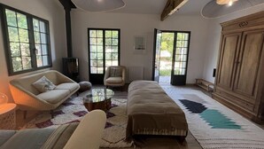 Un salón amplio con sofá, sillón y reposapiés, ventanas grandes y un mueble de madera.