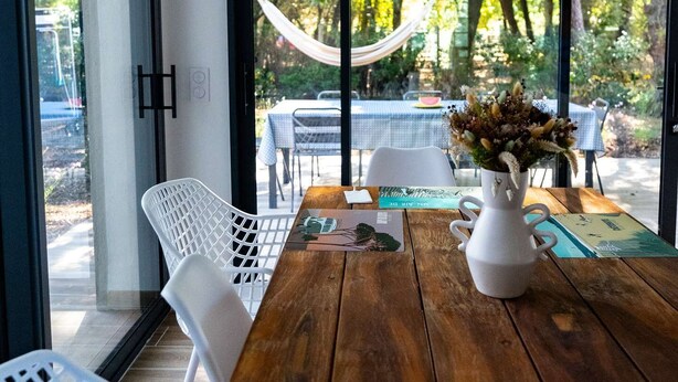 Una mesa de madera con un jarrón blanco, sillas blancas y vistas a un espacio exterior con una hamaca y follaje.