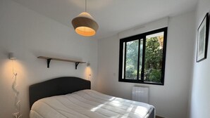 Un dormitorio con cama, lámpara colgante, ventana con vistas a la vegetación y un radiador.
