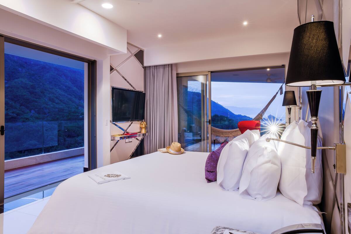 A modern hotel room with a large bed, a flat-screen TV, a desk, and a view of the mountains.