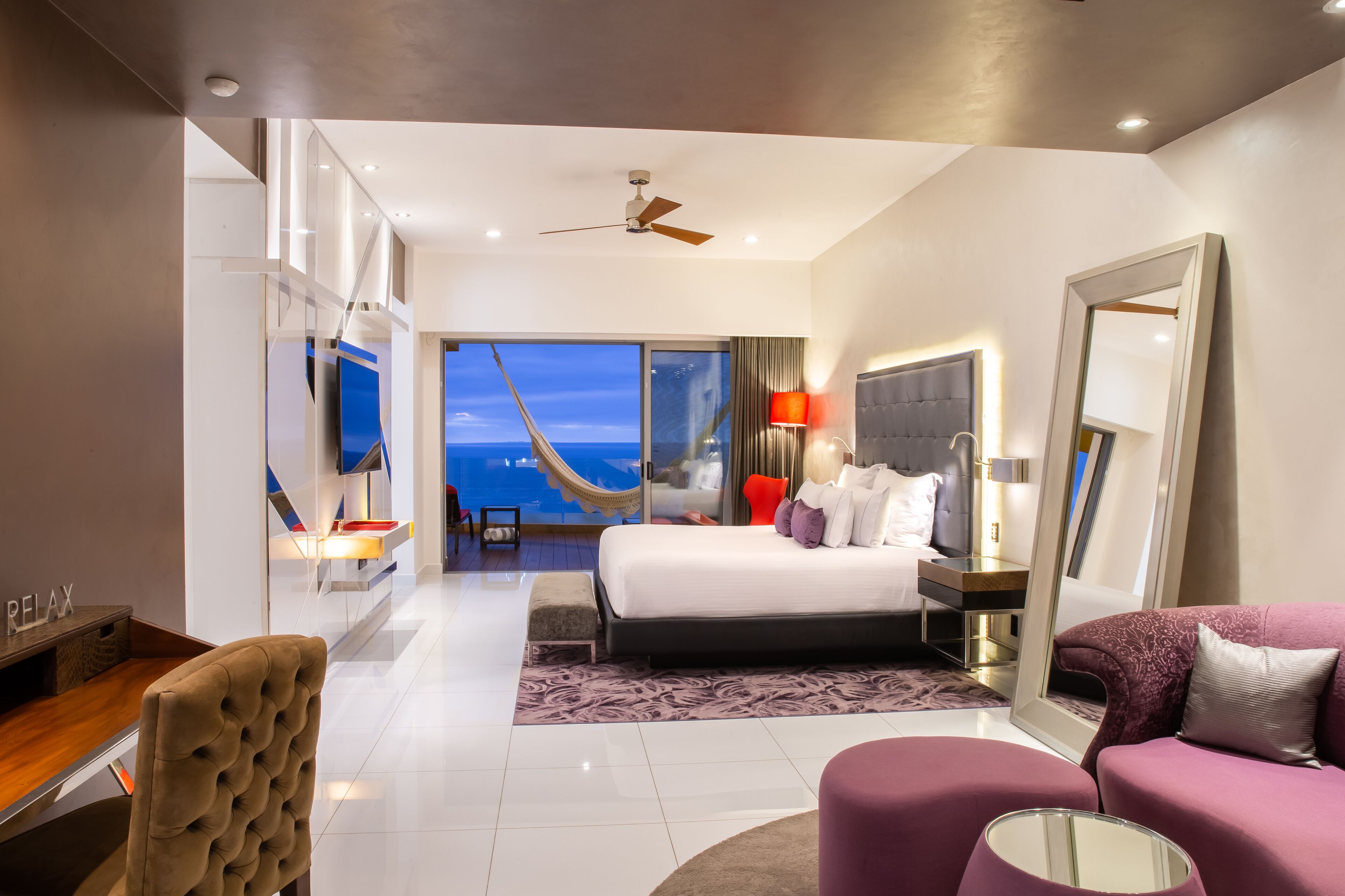 Ein modernes Hotelzimmer mit einem großen Bett, einem Schreibtisch, einem Sessel, einer Couch und einem großen Spiegel. Durch das Fenster ist der Blick auf den Ozean.