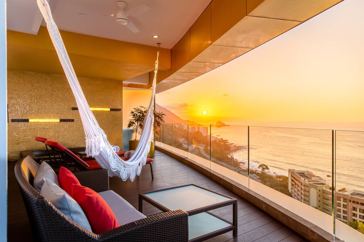 A balcony with a hammock, wicker furniture, and a view of the sunset over the ocean.