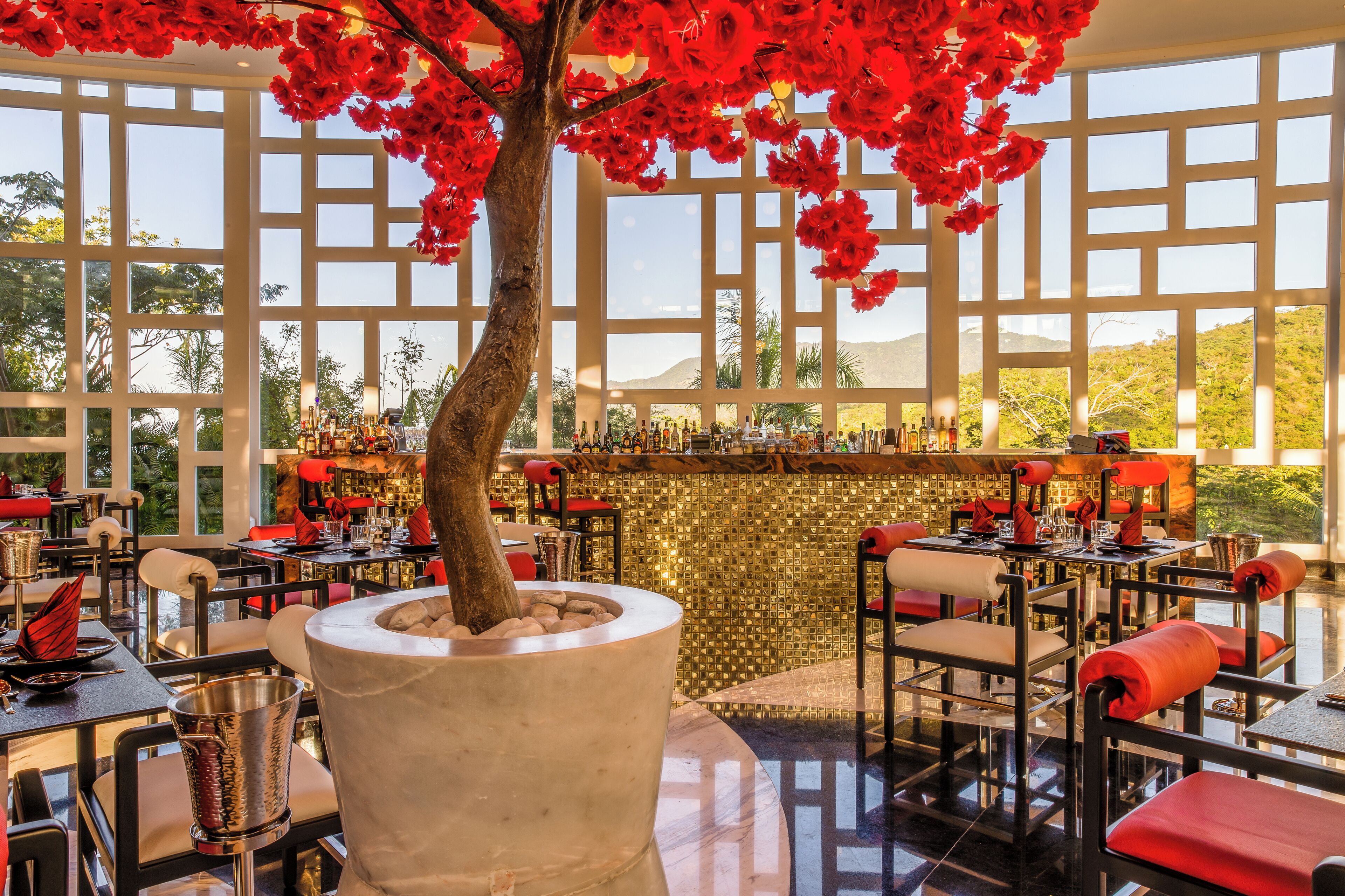 Un restaurante con un gran árbol como centro, mesas con cojines rojos y una zona de bar con botellas.