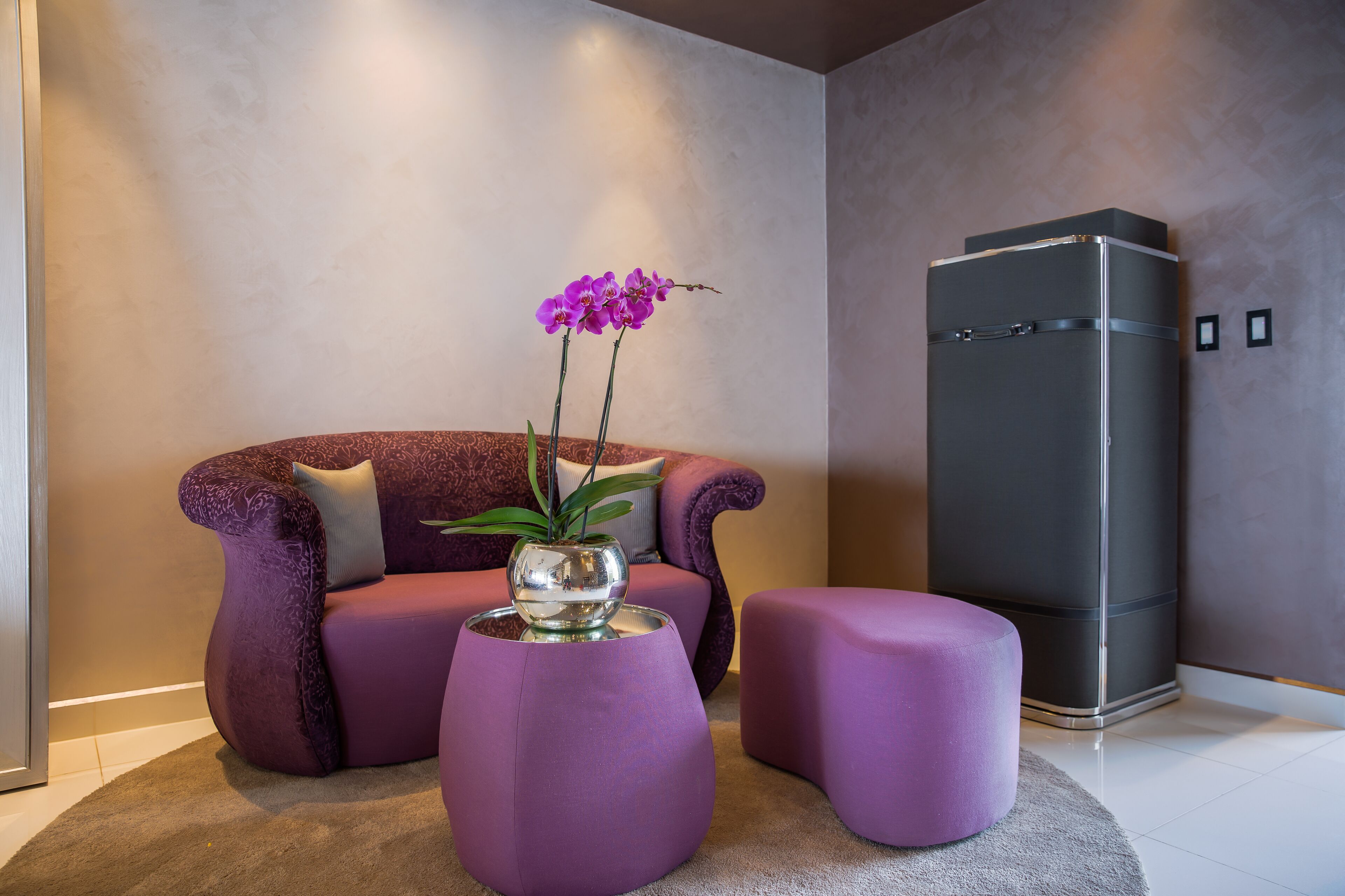 A modern living room with a purple sofa, a glass-top coffee table, and a small ottoman. A tall, dark-colored refrigerator is against the wall.