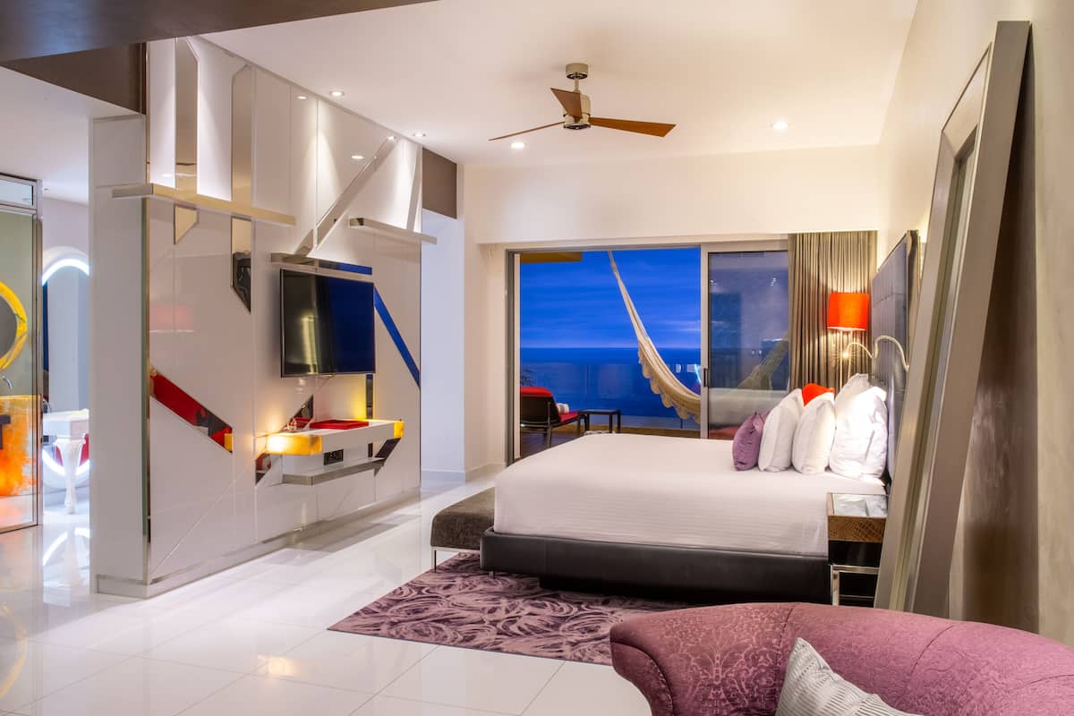 A modern hotel room with a large bed, a TV mounted on the wall, a balcony with a view, and a ceiling fan.