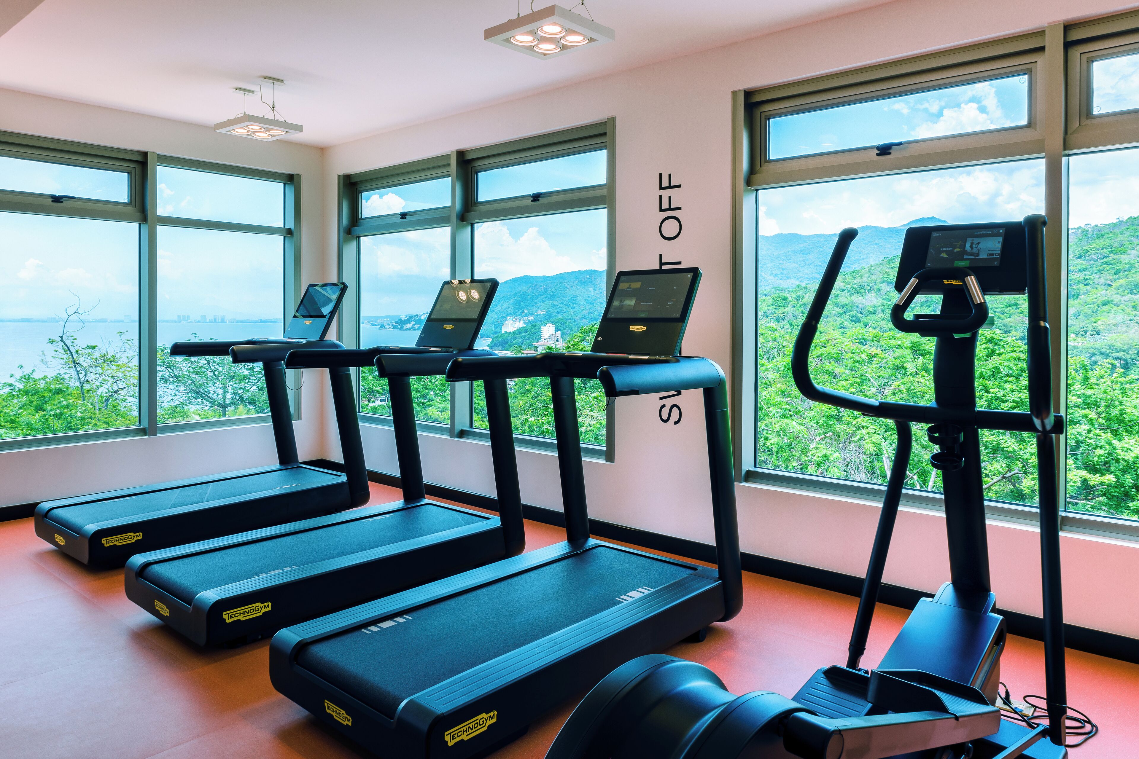 Een sportschool met loopbanden, hometrainers en een roeimachine, grote ramen met uitzicht op groen en bergen, en een bord waarop 