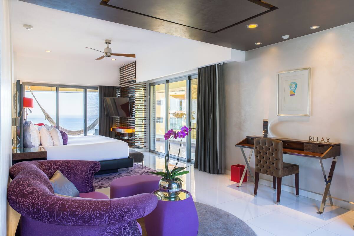 A modern hotel room with a bed, purple seating, a desk, and a view of the ocean.
