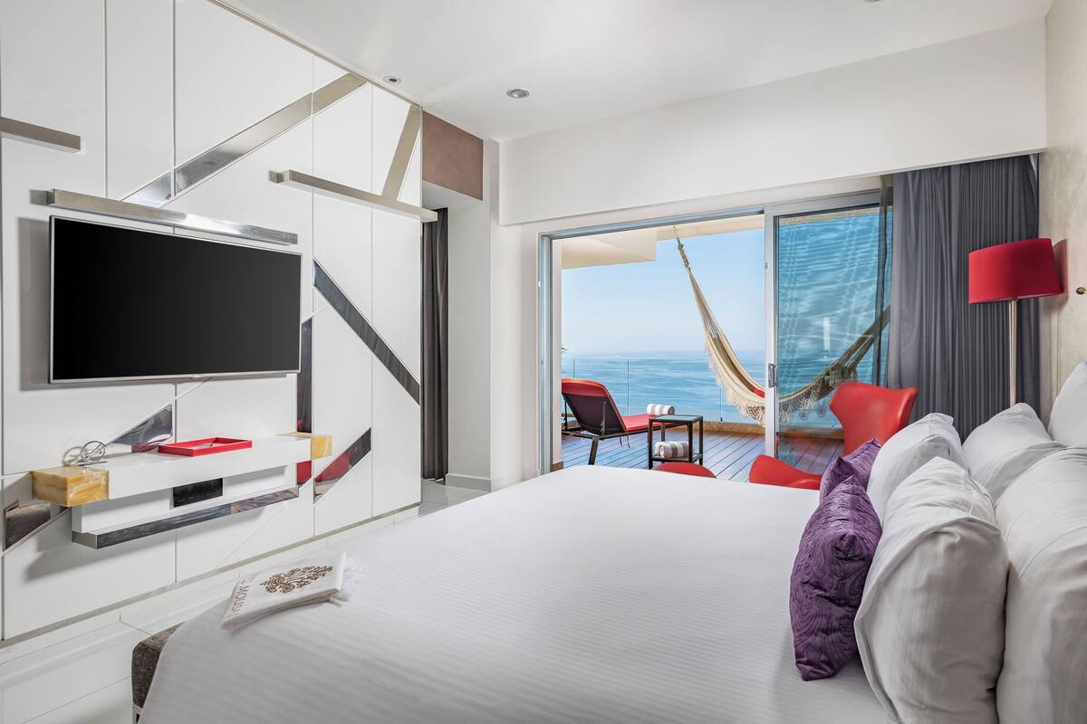 A modern hotel room with a large bed, a TV, a balcony with a hammock, and a view of the sea.