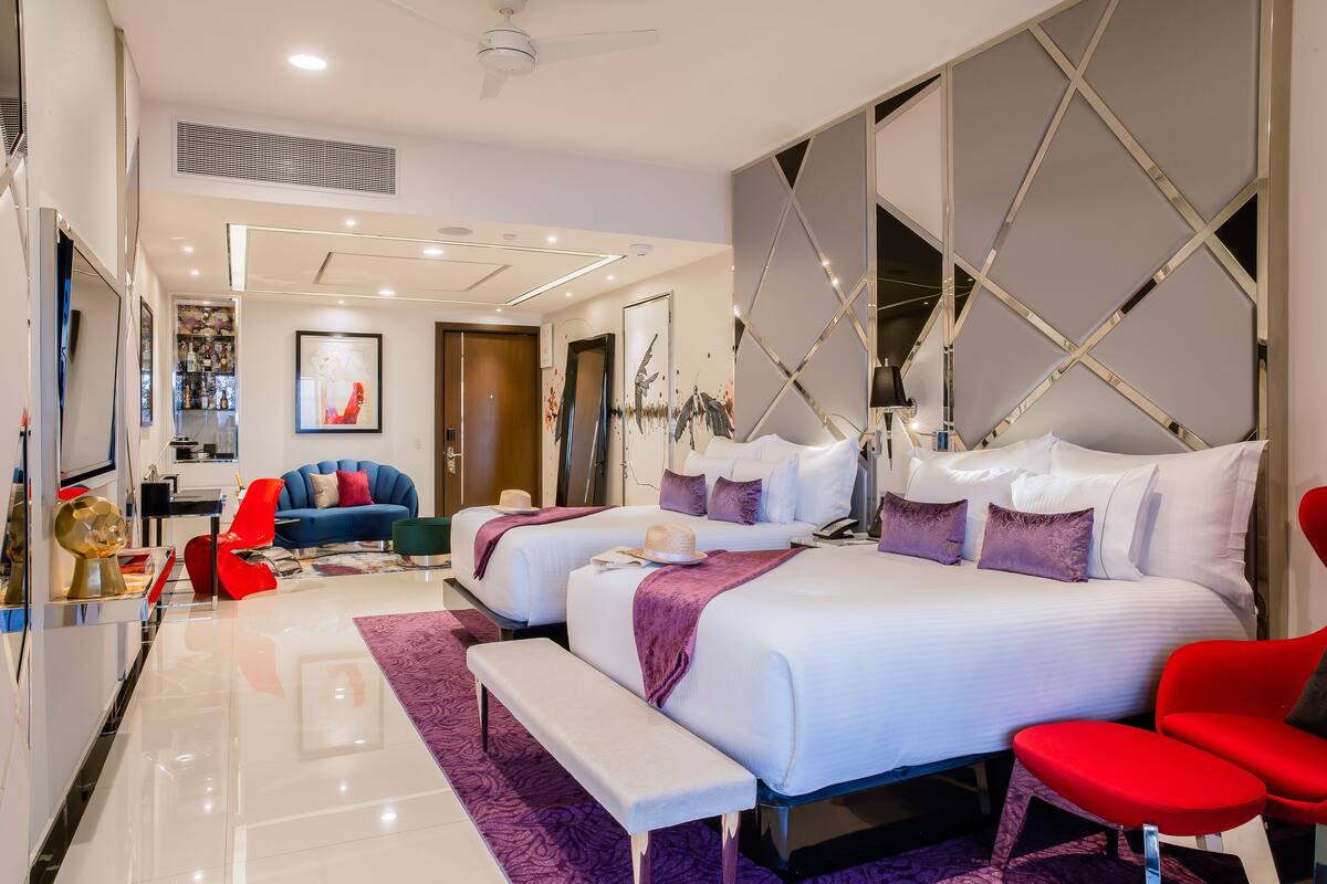 A modern hotel room with two beds, a red chair, a bench, and a large mirror.