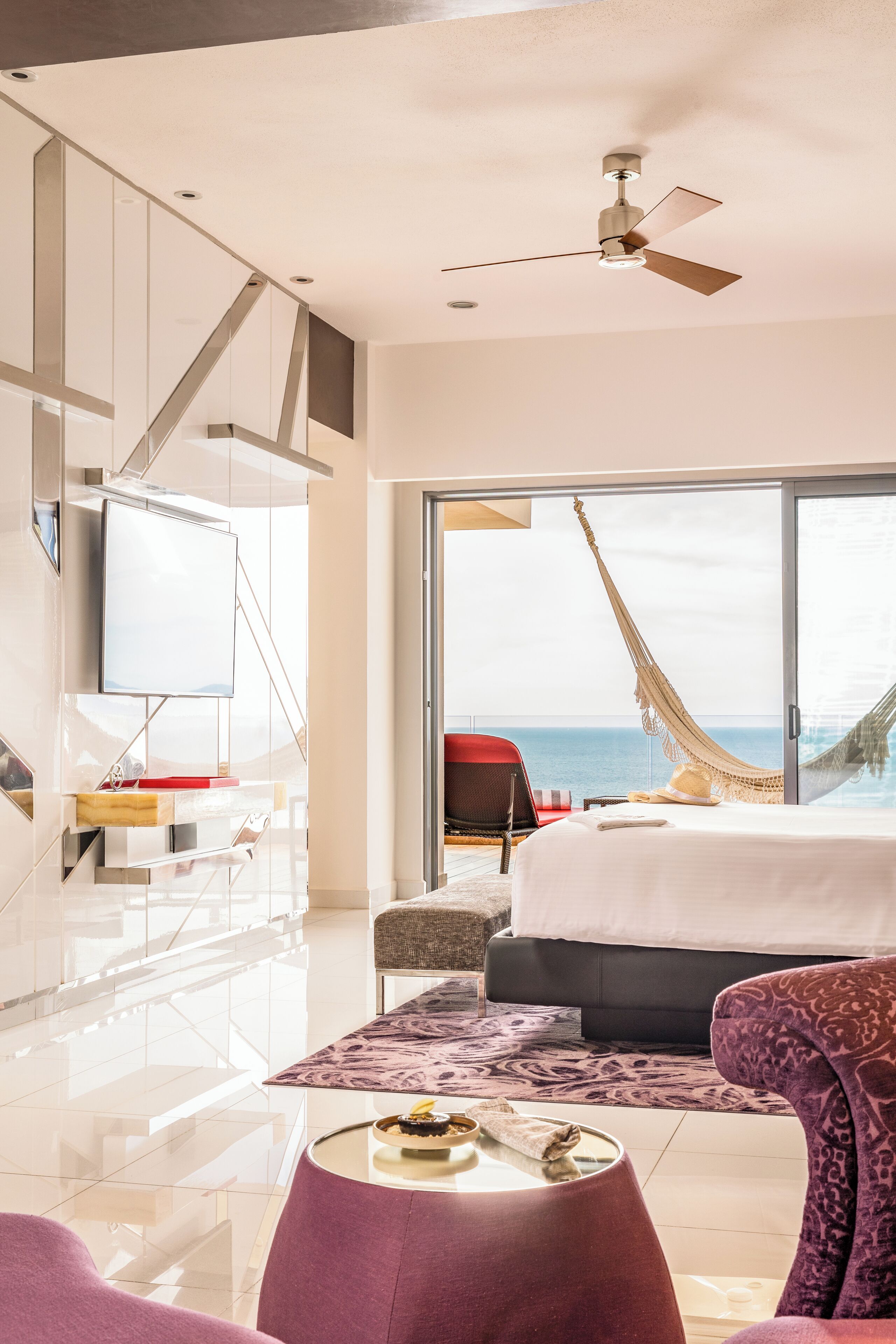 A modern hotel room with a large bed, a TV, a desk, and a view of the ocean.