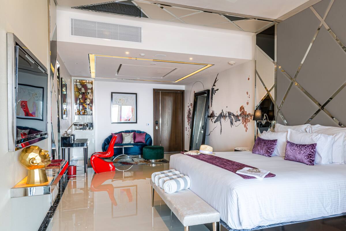 A modern hotel room with a large bed, a red chair, a bench, a desk, and a mirror.