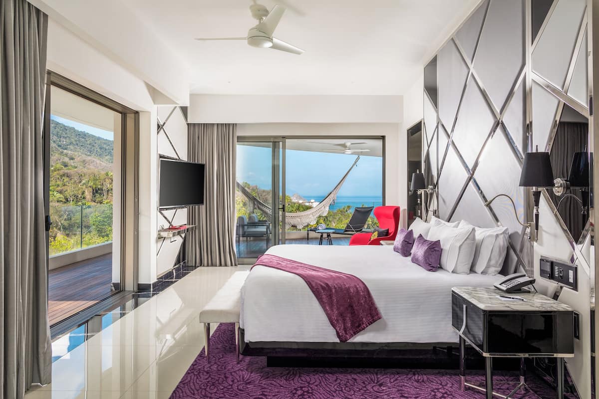 A modern hotel room with a large bed, a TV, a desk, and a view of the ocean.