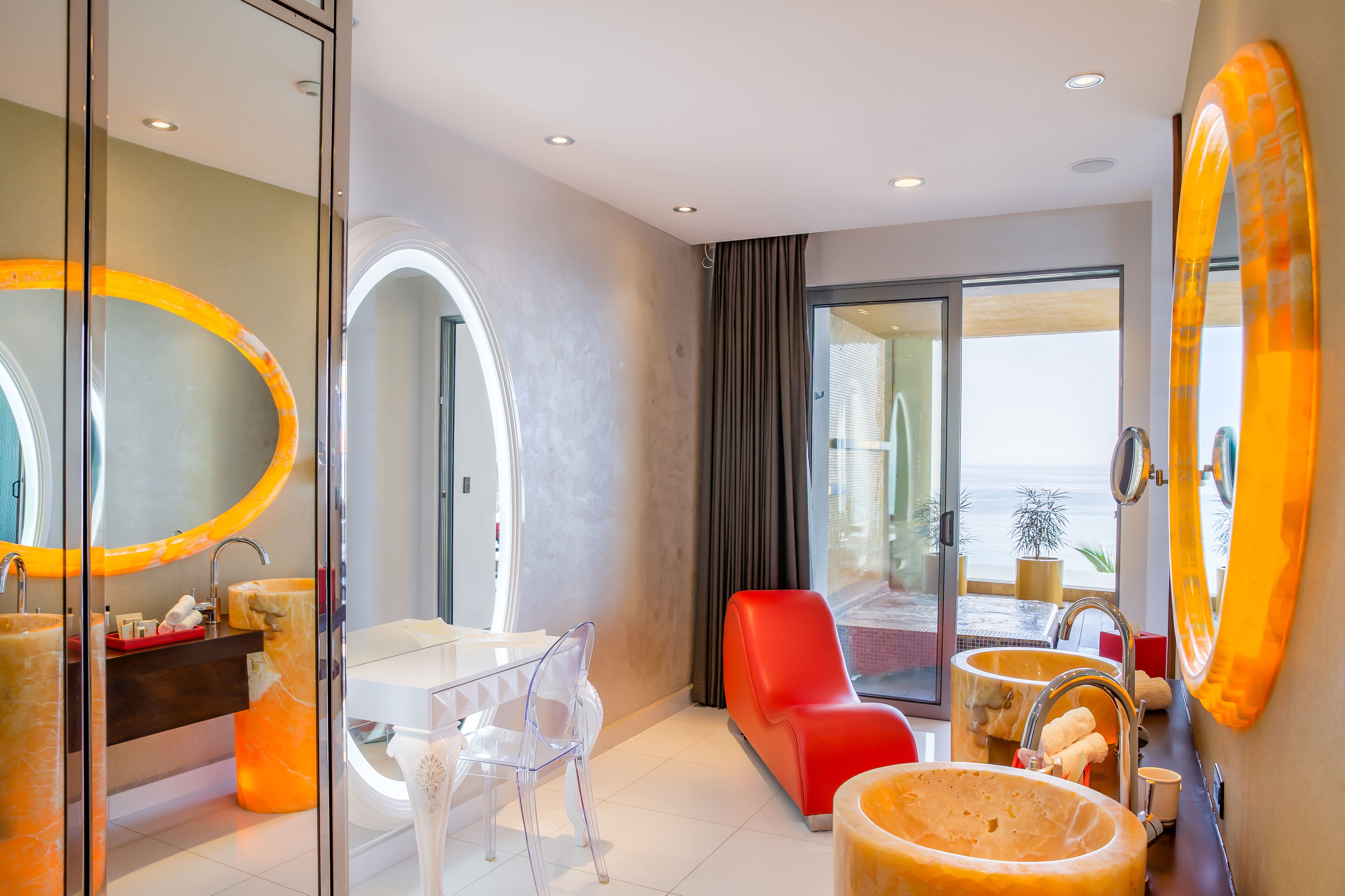 A modern bathroom with a large mirror, a red chair, and a view of the sea.