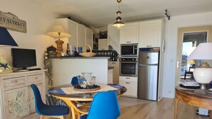 A modern kitchen with white cabinets, a built-in oven, and a stainless steel refrigerator. There is a dining table with blue chairs and a decorative vase. - A Louer T2 Pour Trois Avec Piscine Dans la Résidence (ARZON)