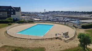 A swimming pool with a sandy deck, surrounded by a fence, and a view of a marina with boats in the background. - A Louer T2 Pour Trois Avec Piscine Dans la Résidence (ARZON)