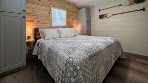 Une chambre avec un plafond en bois, un lit avec une literie à motifs, une table de chevet avec une lampe et une fenêtre avec des stores.