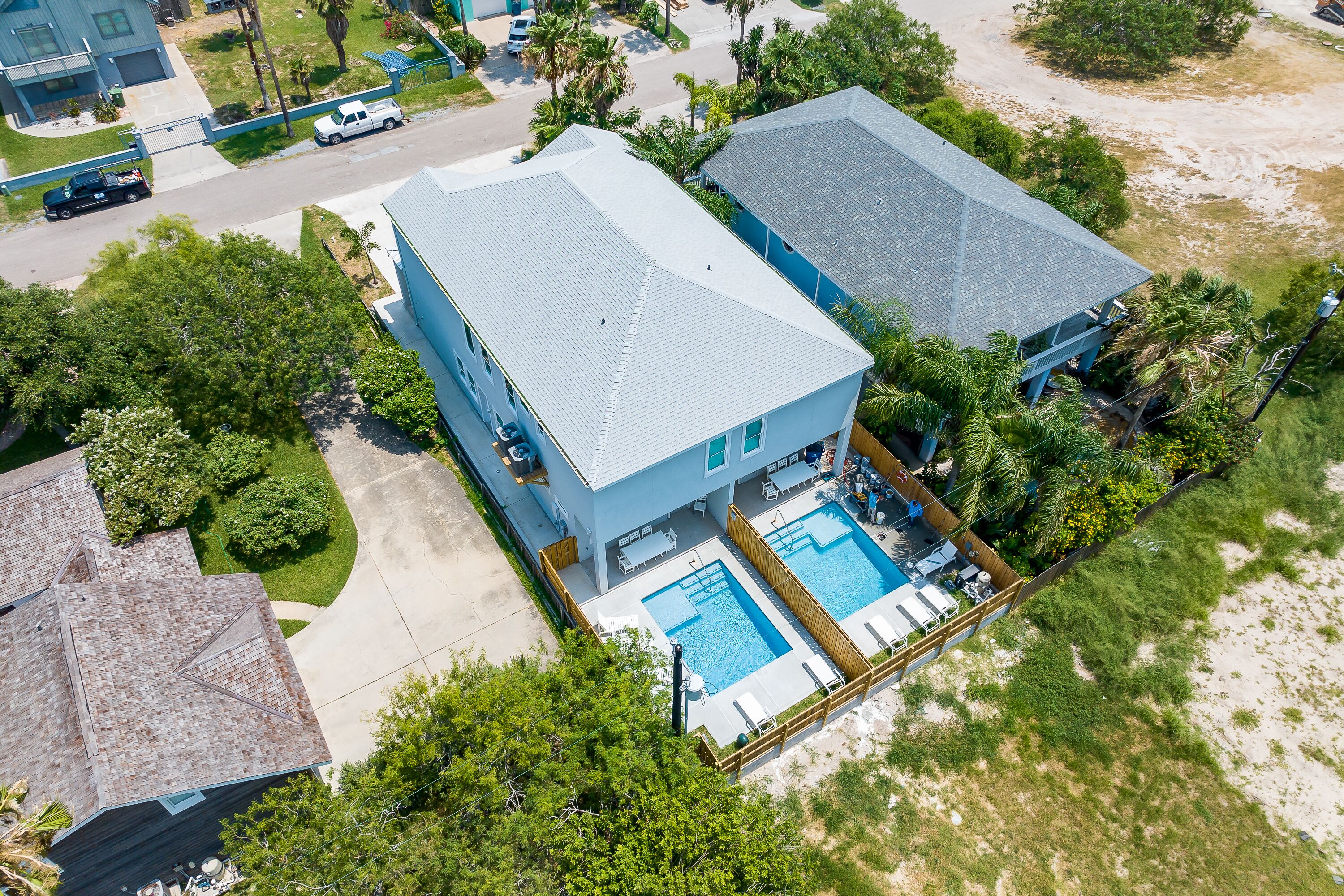 A two-story house with a swimming pool, surrounded by trees and a sandy area.