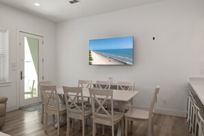Ein Essbereich mit Tisch und Stühlen, einem an der Wand montierten Fernseher und Blick auf einen Strand durch das Fenster.