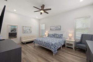Une chambre à coucher comprenant un lit, une chaise, un bureau, un ventilateur de plafond et des œuvres d’art accrochées aux murs.