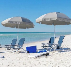 Ein Strand mit zwei Sonnenschirmen, vier Liegestühlen und einer Kühlbox im Sand.