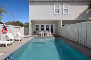 Ein Swimmingpool mit gefliestem Rand, umgeben von einem weißen Zaun, und eine Terrasse mit weißen Stühlen und einem Tisch.