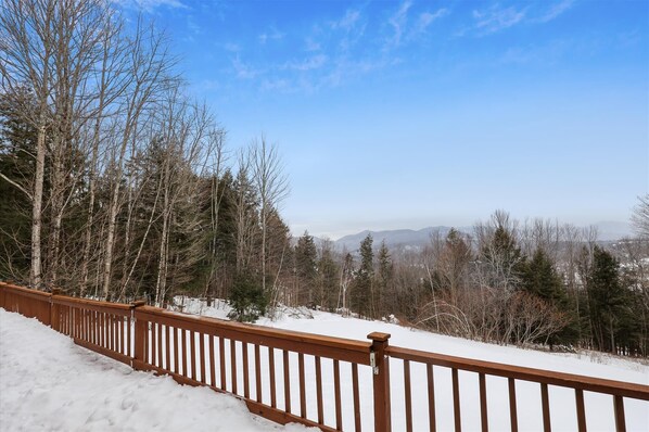 A snow-covered deck with a wooden railing overlooking a forest and distant mountains. - Dug's House: 4 br, pet friendly, deck w/ Mansfield views! (Stowe)