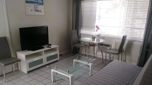 A modern living room with a flat-screen TV, a glass coffee table, a sofa, and a dining area with a small table and chairs.