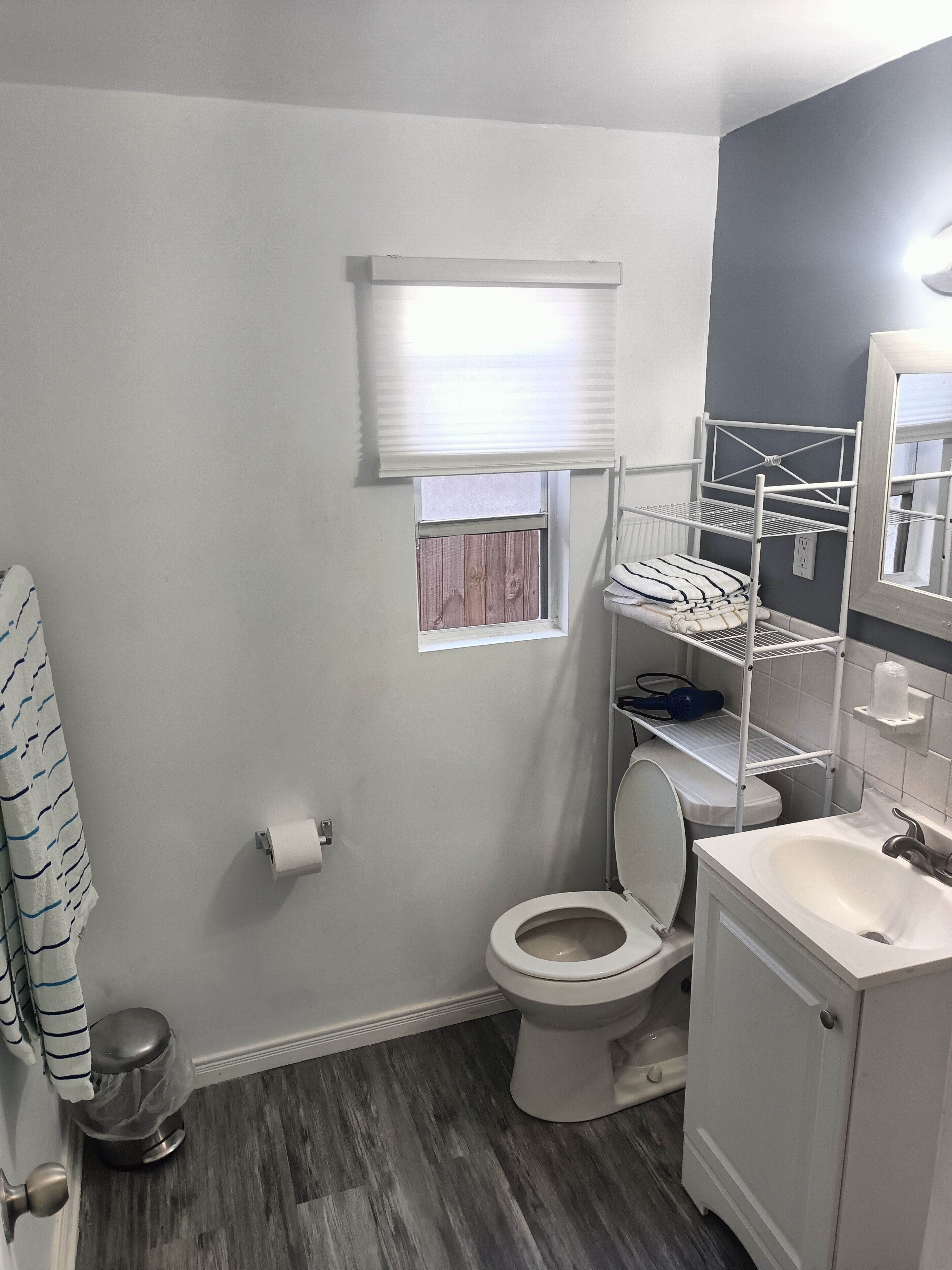 A bathroom with a toilet, a sink, a window with blinds, a trash can, and a towel rack.