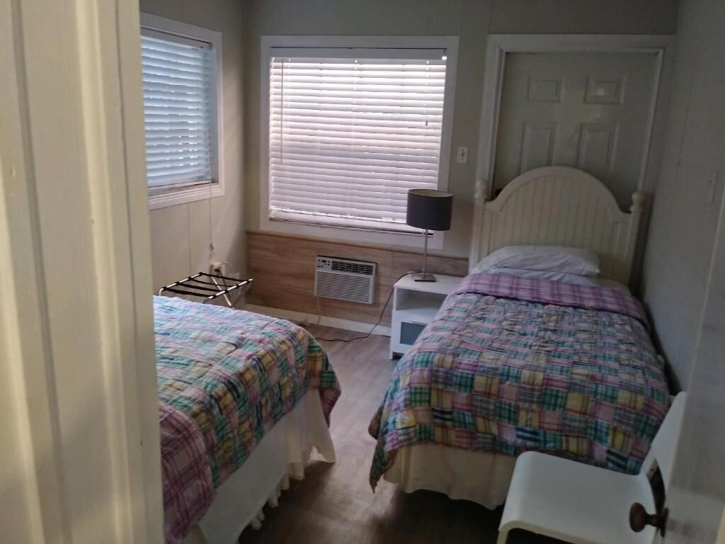Ein Schlafzimmer mit zwei Betten, einem Fenster mit Jalousien, einem Nachttisch mit Lampe und einer Tür.