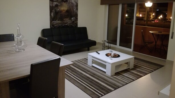 A modern living room with a black sofa, a dining table with chairs, a white coffee table, and a large Buddha painting on the wall. - T2 Novo Alto do Quintão (Portimão)