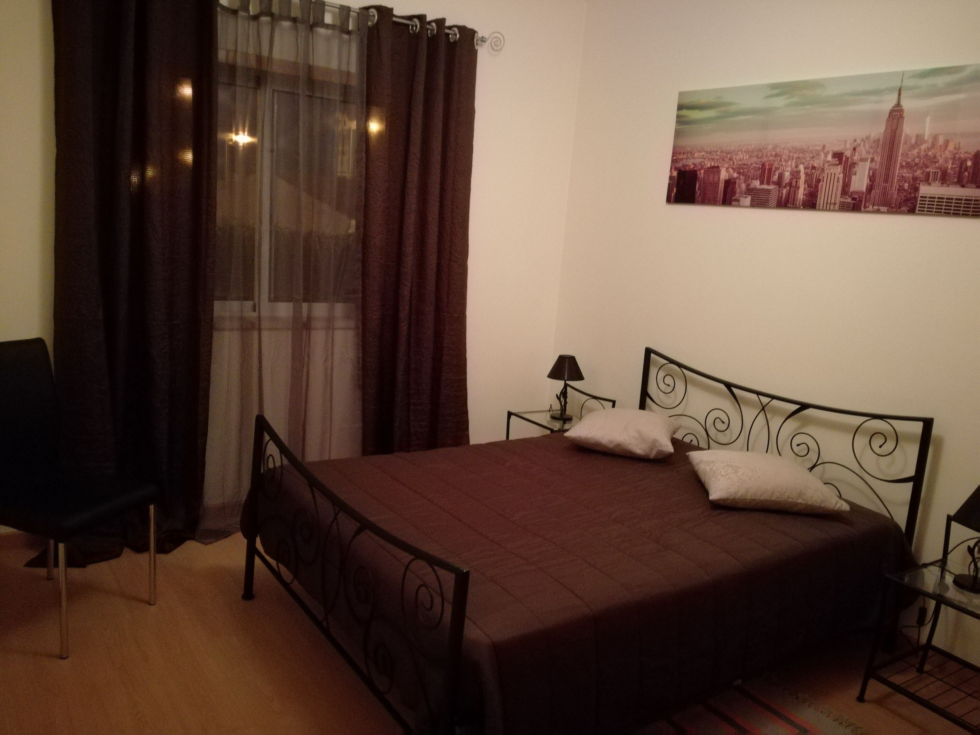 Ein Schlafzimmer mit Bett, einem Sessel, einem Nachttisch, einer Lampe und einem Stadtbild an der Wand.