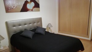A bedroom with a tufted headboard, a black bedspread, two pillows, a bedside table with a lamp, and a wooden wardrobe. - T2 Novo Alto do Quintão (Portimão)