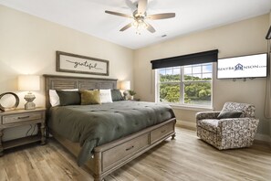 A bedroom with a bed, nightstands, a chair, a ceiling fan, a window with a view, and a wall-mounted TV. - Lakefront on Taneycomo with Fireplace, Just Minutes from Branson Landing (Branson)