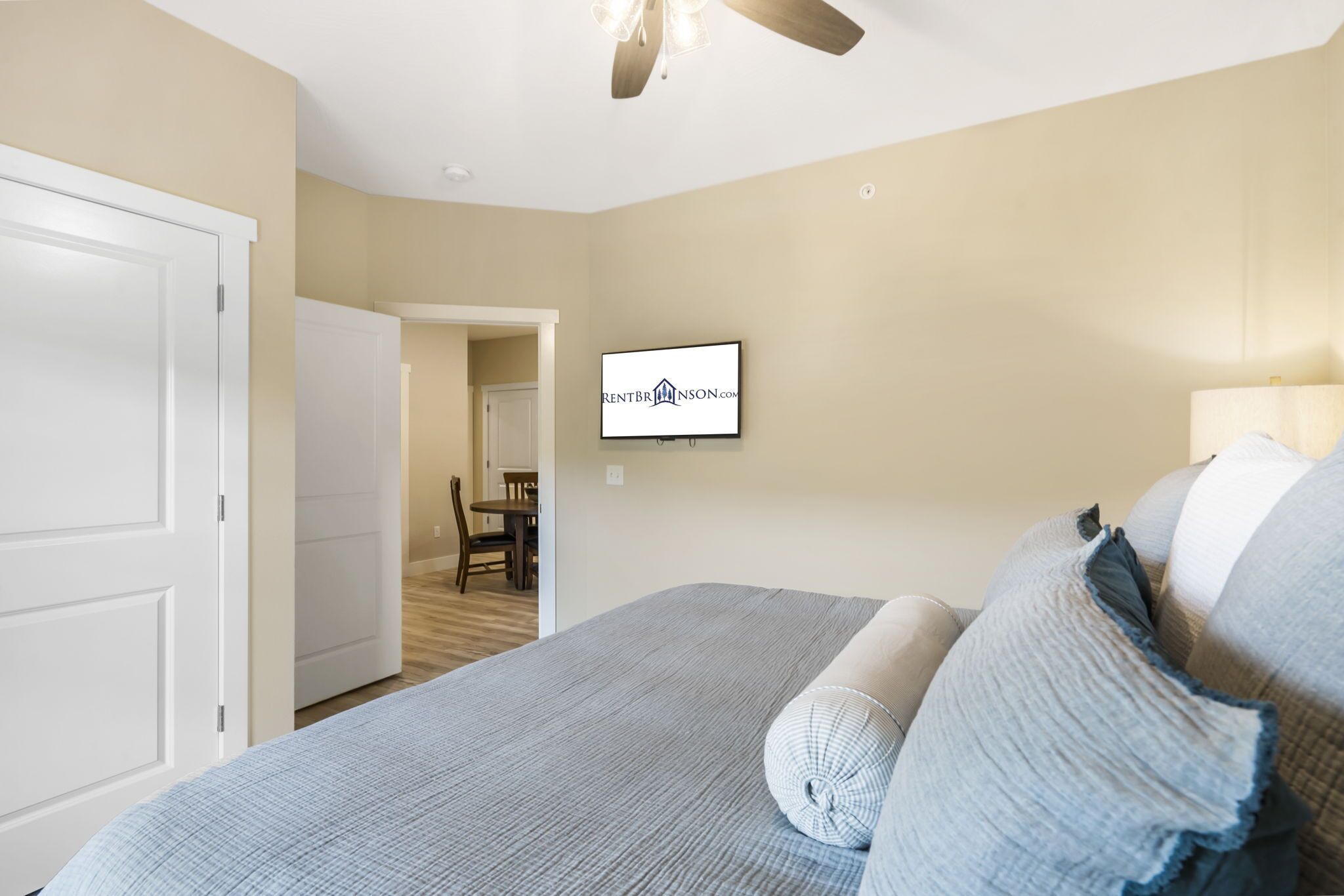 A bedroom with a bed, a ceiling fan, a TV mounted on the wall, and a door leading to another room.