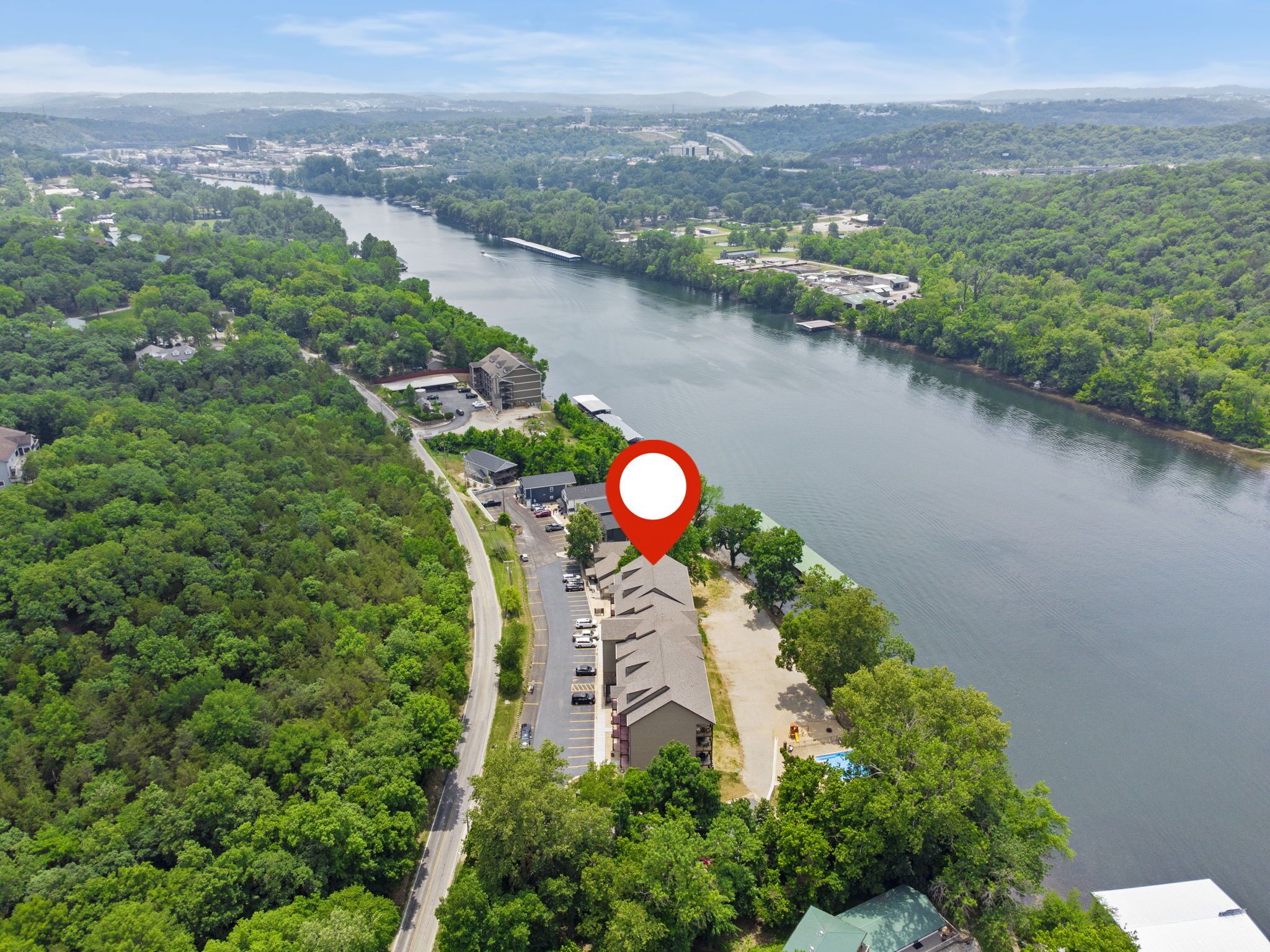 An aerial view of a riverside property with a parking lot, buildings, and dense greenery.