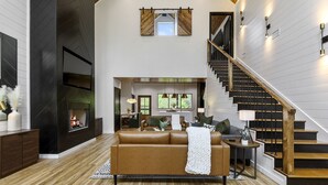 A spacious living area with a fireplace, a brown leather sofa, a staircase, and a modern kitchen in the background. - Play-Set, Darts, Large Back Deck, Hot Tub, Pets, Hidden Jewel (Broken Bow)