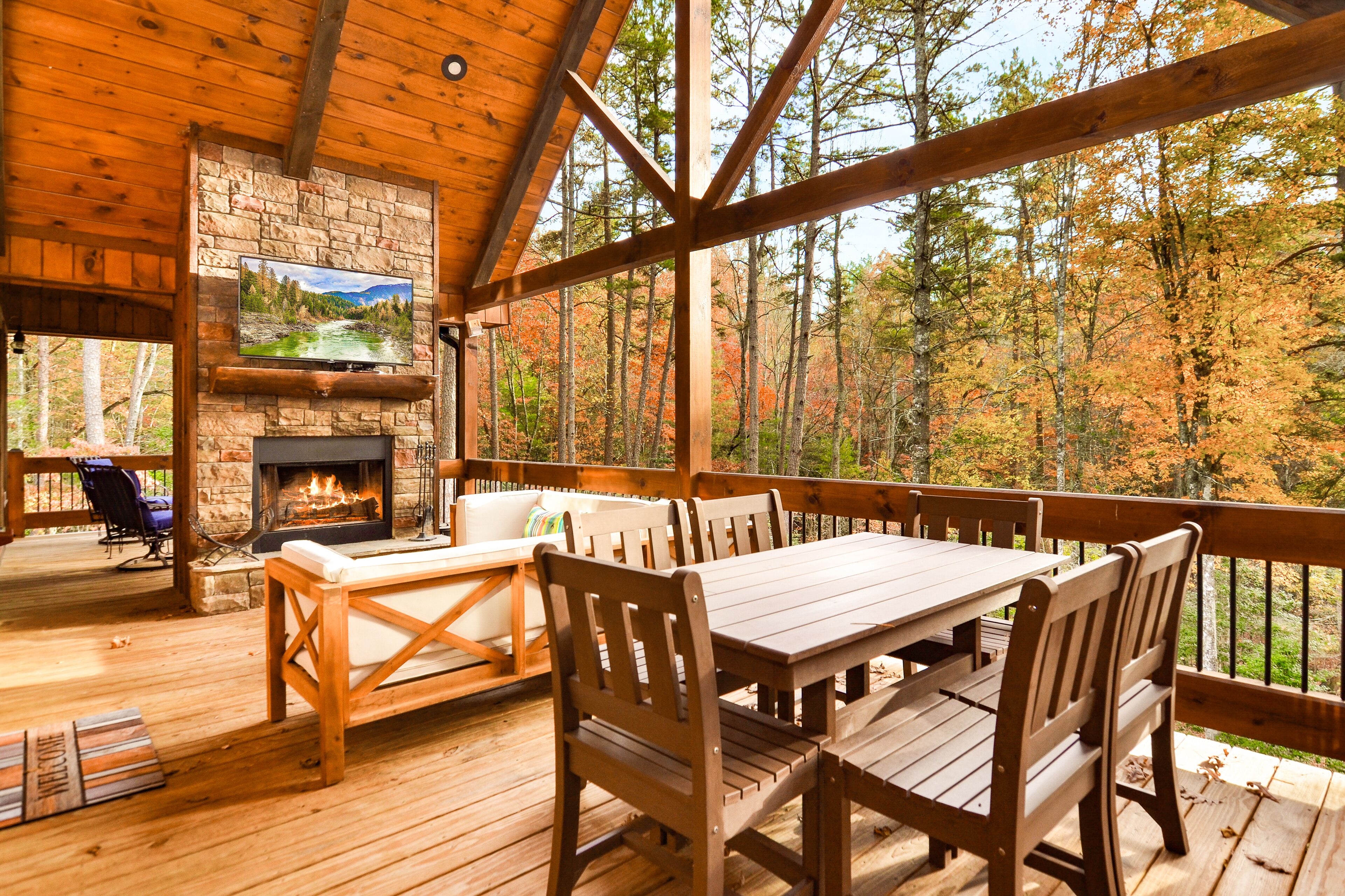 A wooden deck with a stone fireplace, a TV mounted on the wall, and a view of autumn trees.