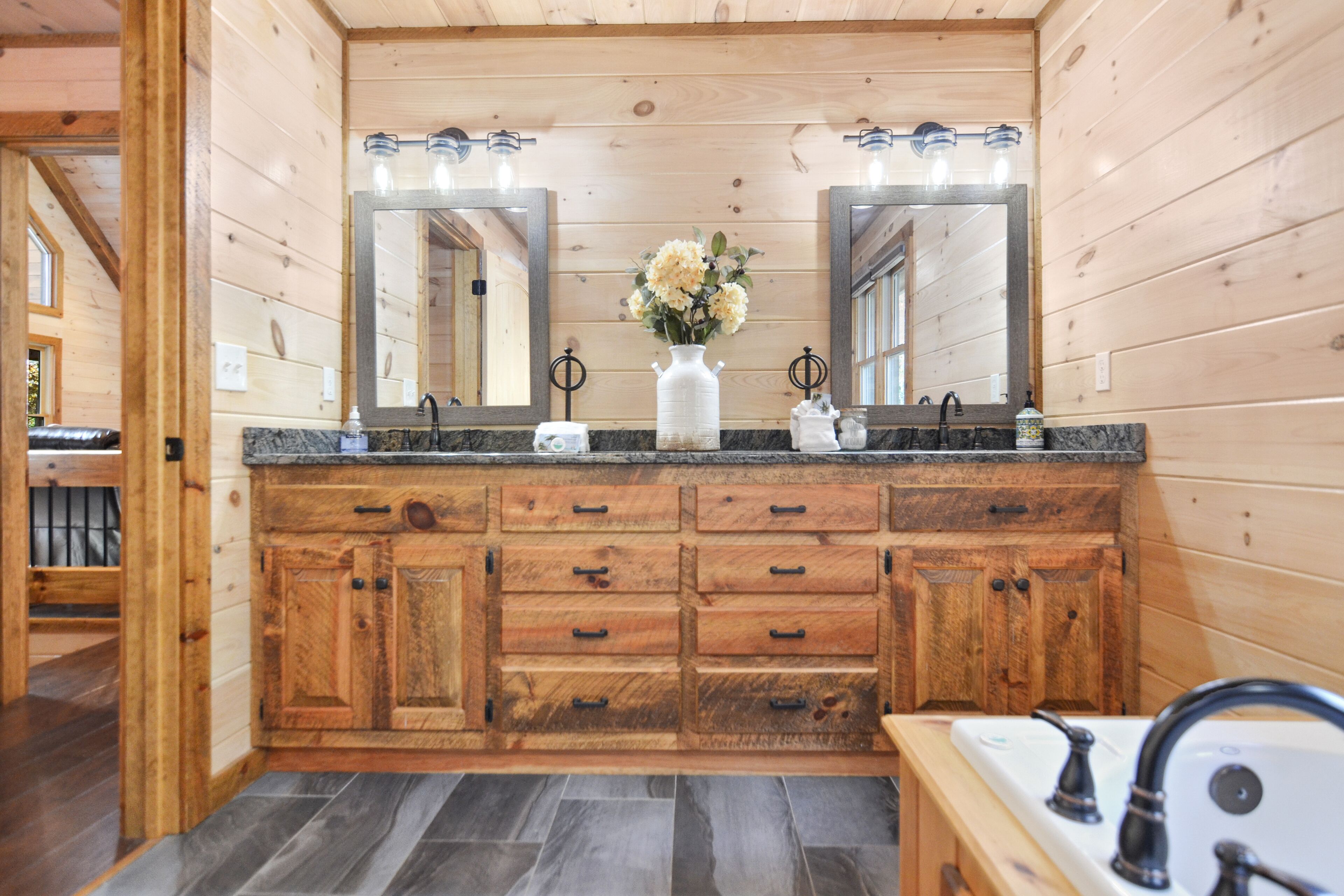Une salle de bain de style rustique, avec des murs en bois, un grand meuble-lavabo, deux miroirs et un vase de fleurs.