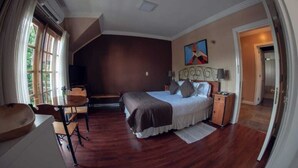 A bedroom with a large bed, a desk, and a chair. There is a window with curtains, a painting on the wall, and a door leading to another room.