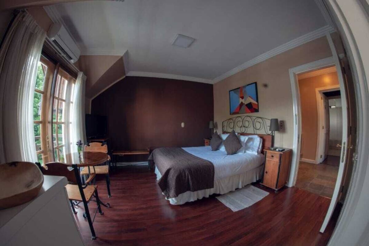 Quarto com cama grande, escrivaninha e cadeira. Há uma janela com cortinas, um quadro na parede e uma porta que dá para outro cômodo.