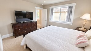 Un dormitorio con cama, cómoda, televisor, ventana con cortinas y mesita de noche con lámpara.