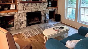 Una sala de estar con chimenea de piedra, estantes de madera, ventilador de techo y ventanas amplias.