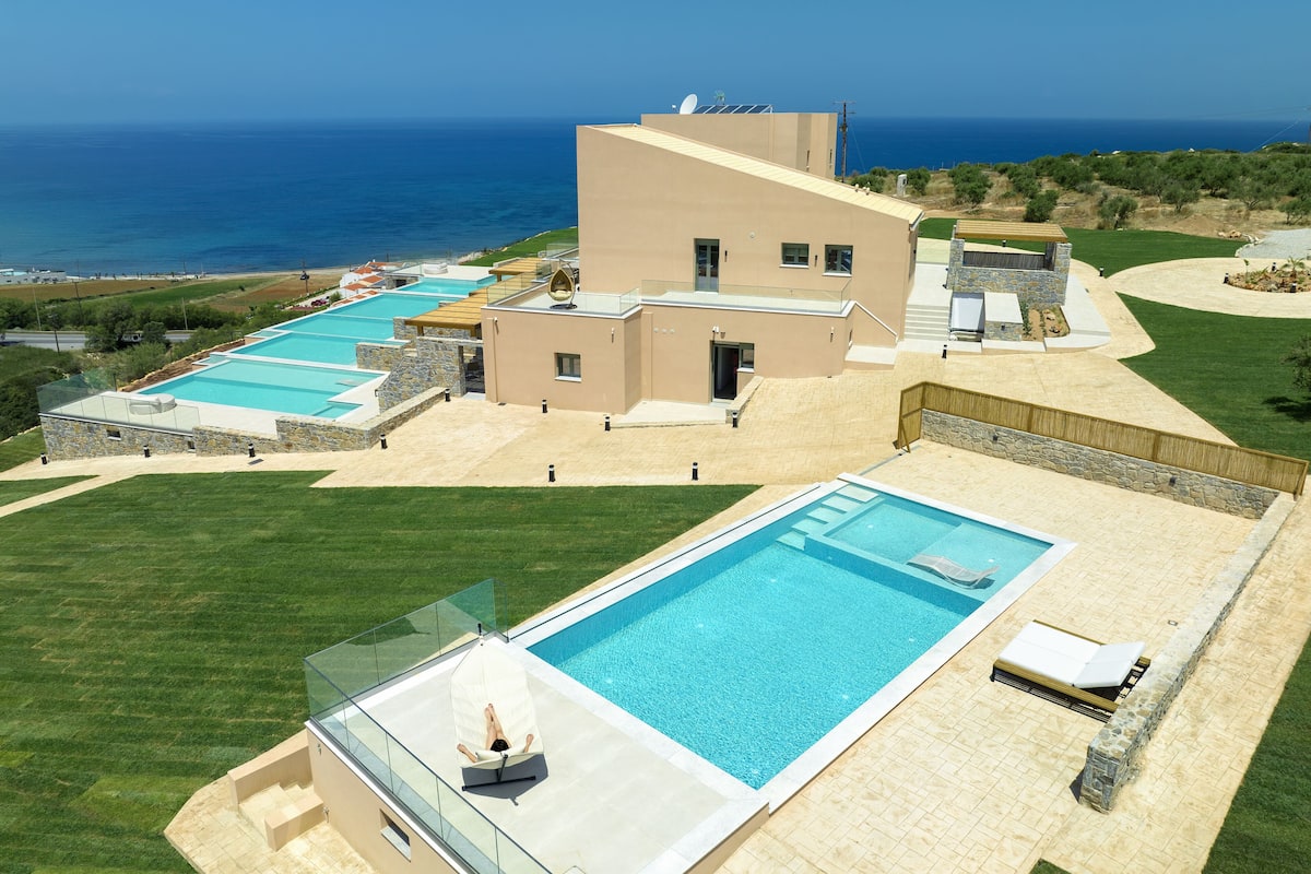 A modern resort with multiple swimming pools, a large central pool, and a view of the ocean.