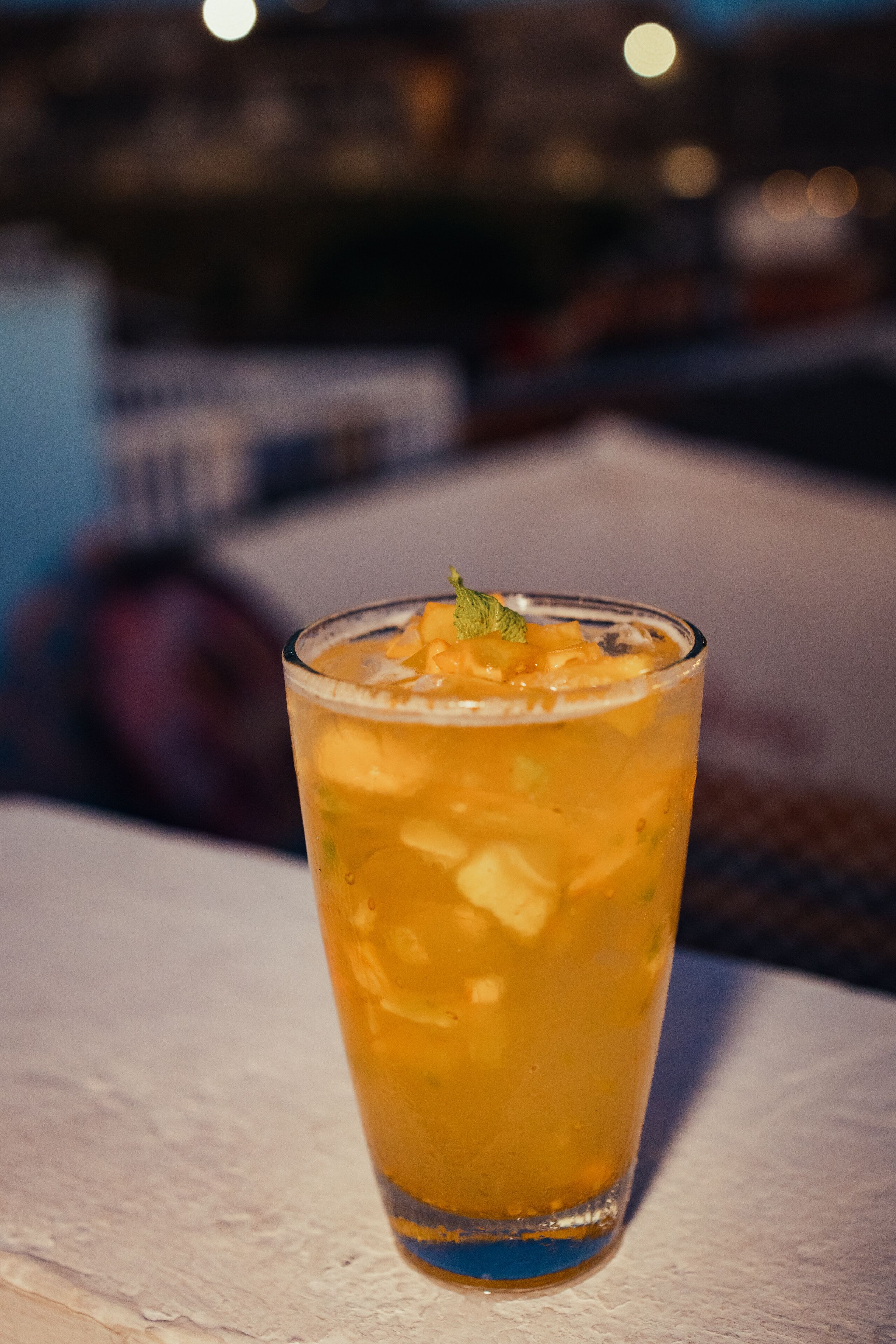 A tall glass with a yellow beverage, ice cubes, and a mint garnish, placed on a white surface.