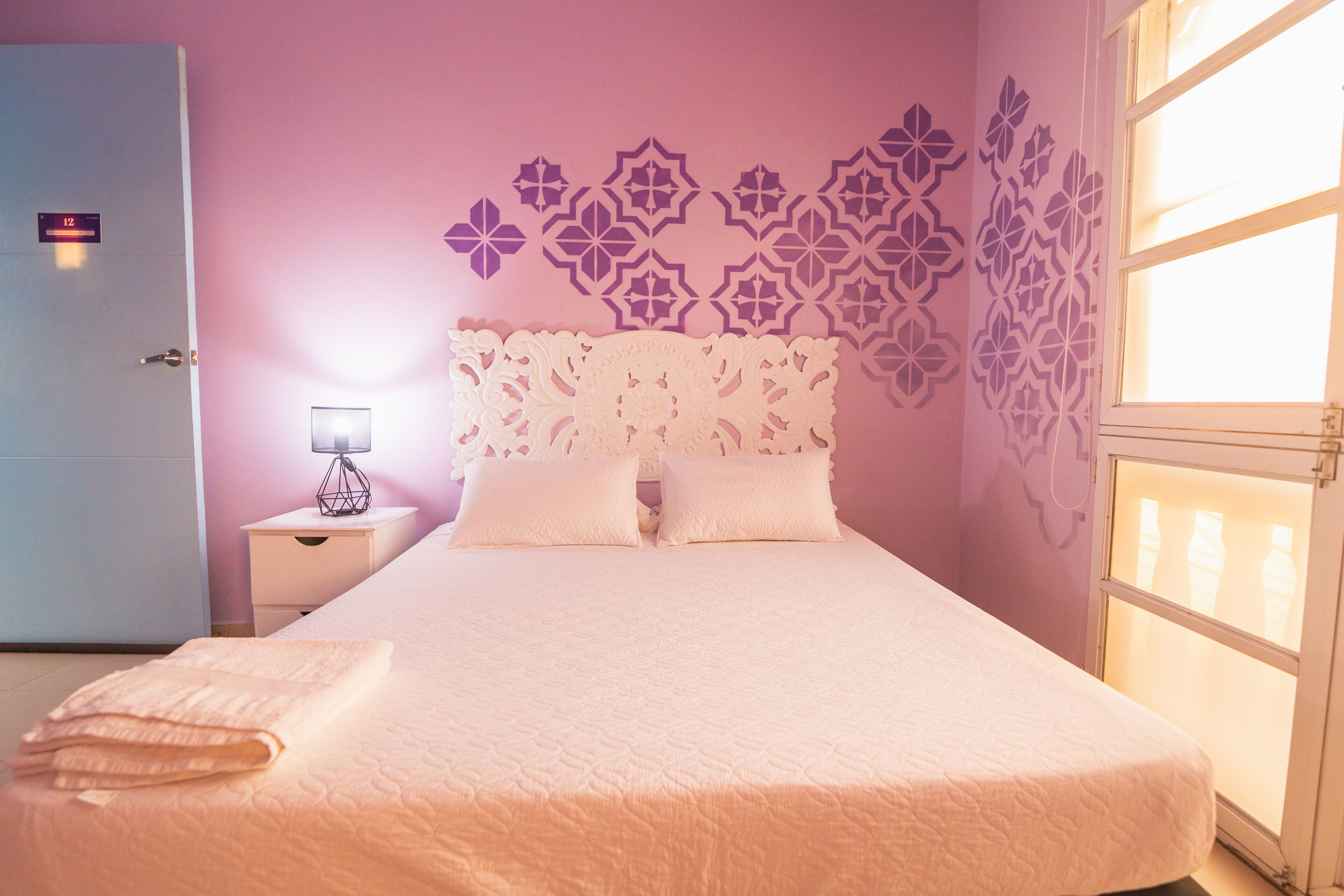 Una cama bien hecha con una colcha blanca y almohadas, una mesita de noche con lámpara, y una pared con diseños decorativos.