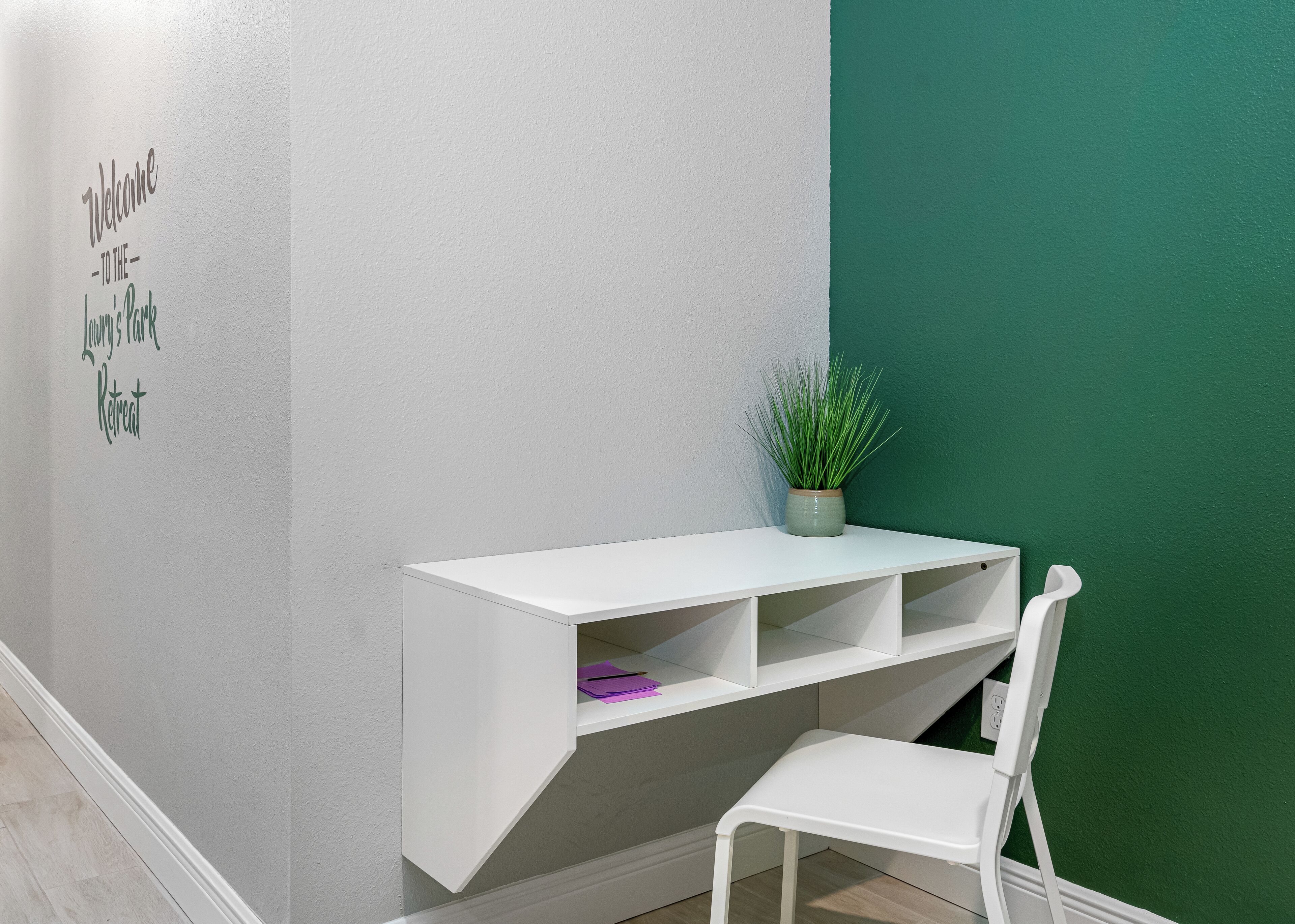 Un bureau blanc avec des étagères, une chaise blanche, une plante en pot et un mur orné d’une pancarte indiquant « Bienvenue chez… ».