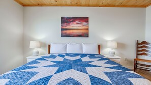 A bedroom with a large bed, two bedside tables with lamps, a wooden headboard, and a painting of a sunset on the wall. - Olver Farm Cottage - Newly built cottage only feet from the shores of Cayuga Lake! (Romulus)
